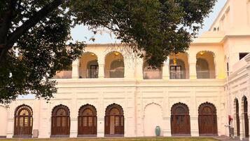 Un edificio con múltiples ventanales arqueados y puertas, un patio con una fuente y un árbol grande al frente.