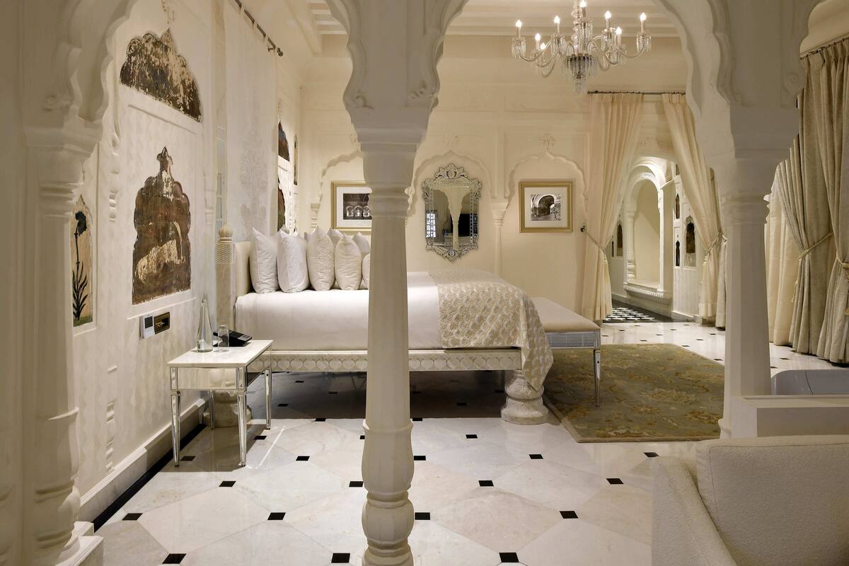 A luxurious bedroom with a large bed, ornate columns, and a chandelier.
