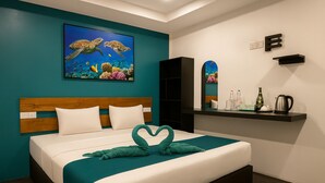 A hotel room with a bed, a teal wall, a painting of sea turtles, a heart-shaped towel, a shelf with toiletries, and a mirror. - Elysian Hideaway Maafushi (Maafushi)