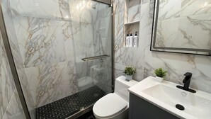 Un baño moderno con ducha de vidrio, un lavabo blanco con grifo negro y paredes de mármol.