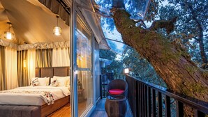 A bedroom with a large bed, a wicker chair, and a balcony with a view of trees. - The Whispering Oaks (Nainital)