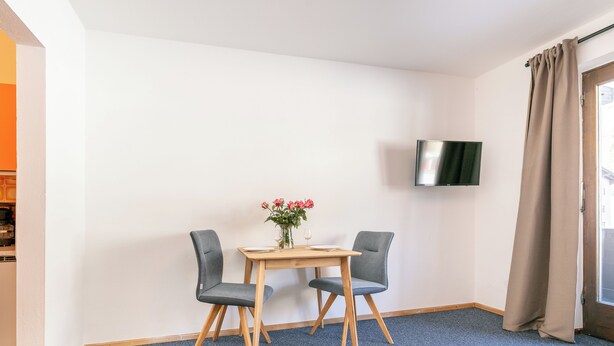 A hotel room with a blue carpet, a wooden table with two chairs, a vase with flowers, and a TV mounted on the wall.