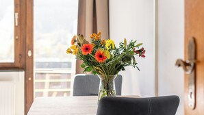 Une table à manger ornée d’un vase de fleurs, une chaise, et une vue sur un bâtiment à travers une fenêtre.