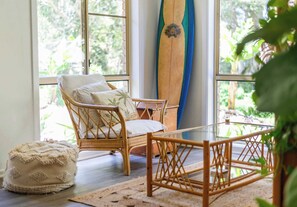 A wicker chair with cushions, a wooden coffee table, a surfboard, and a window with a view of greenery.