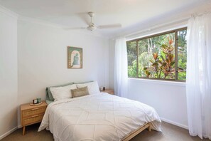 A bedroom with a bed, a nightstand, a ceiling fan, a window with curtains, and a view of greenery outside.