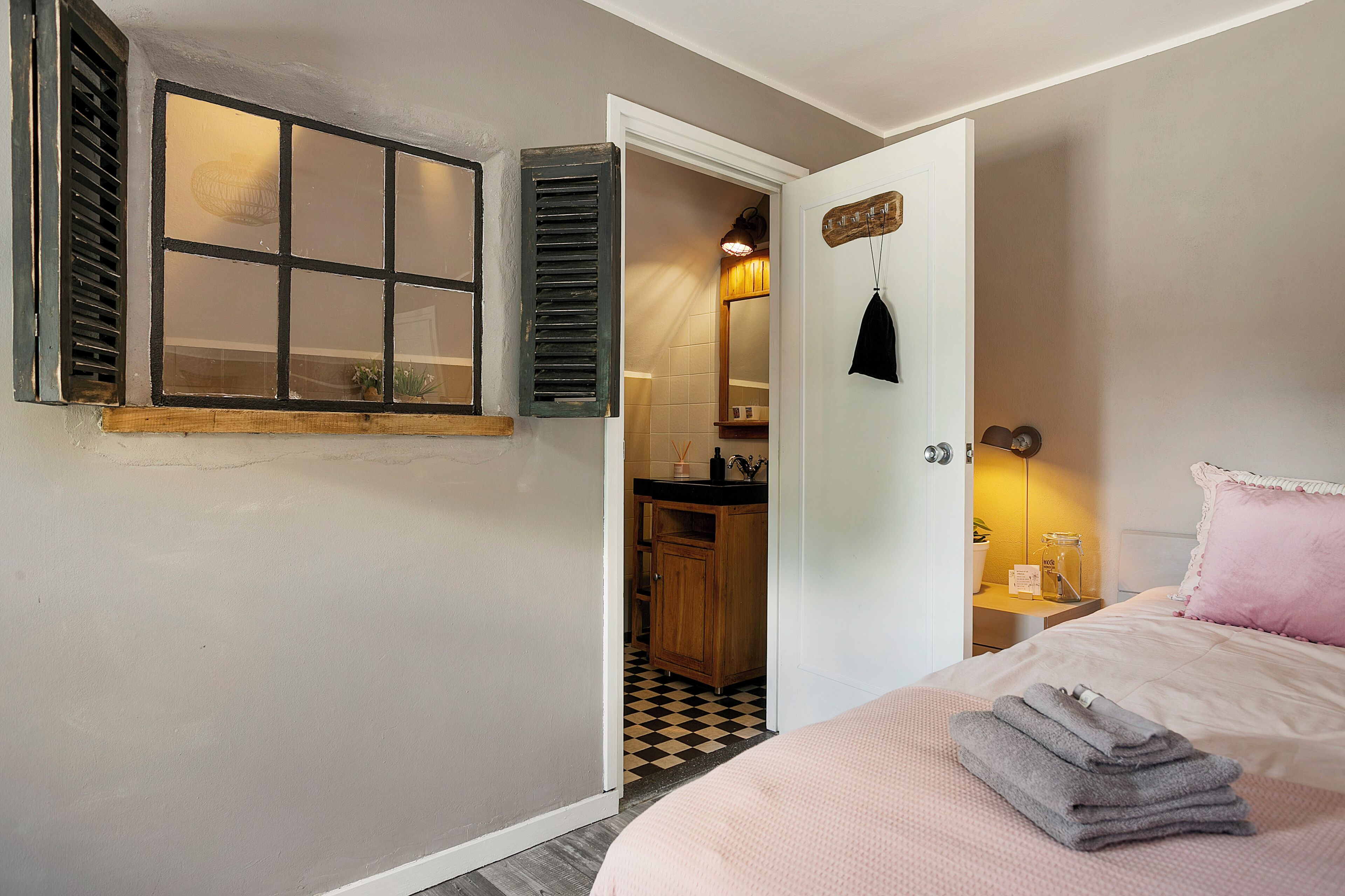 Un dormitorio con cama, mesita de noche, ventana con contraventanas y baño visible a través de una puerta abierta.