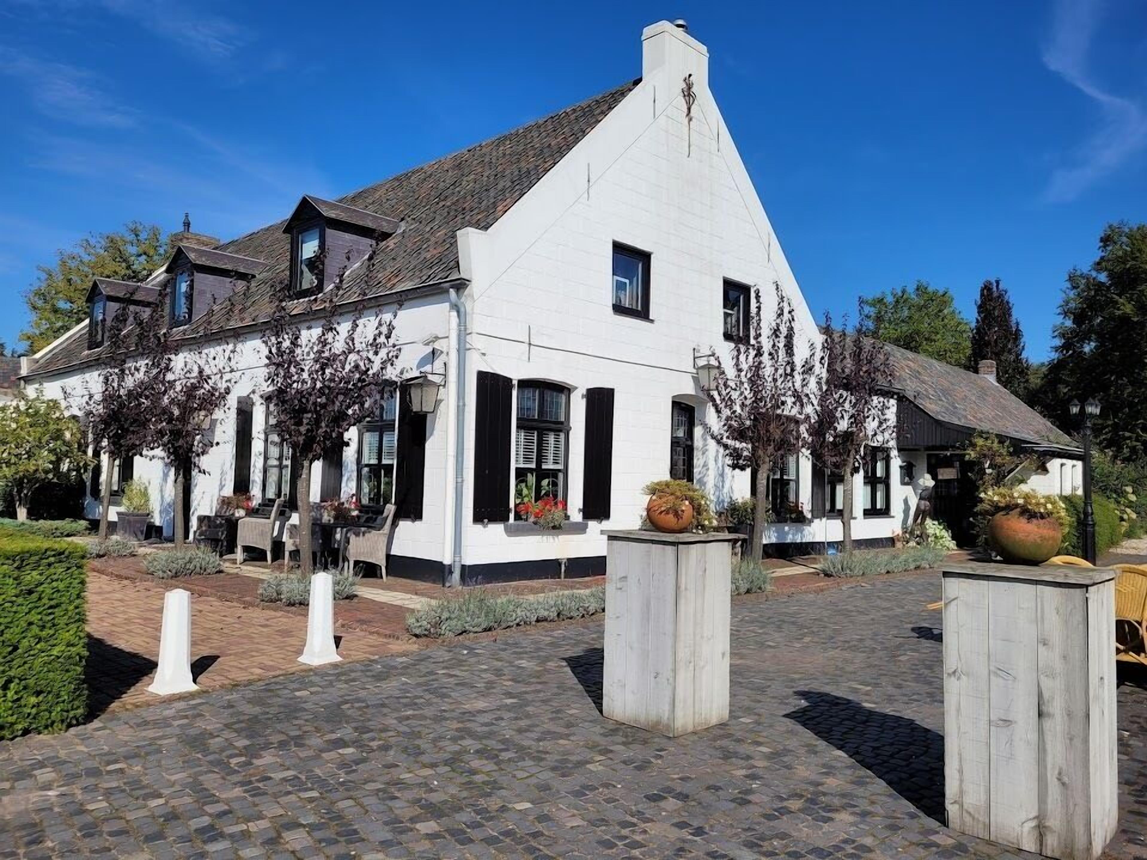 Een wit gebouw met een schuin dak, zwarte luiken, en een bestraat entreegebied met paaltjes en plantenbakken.