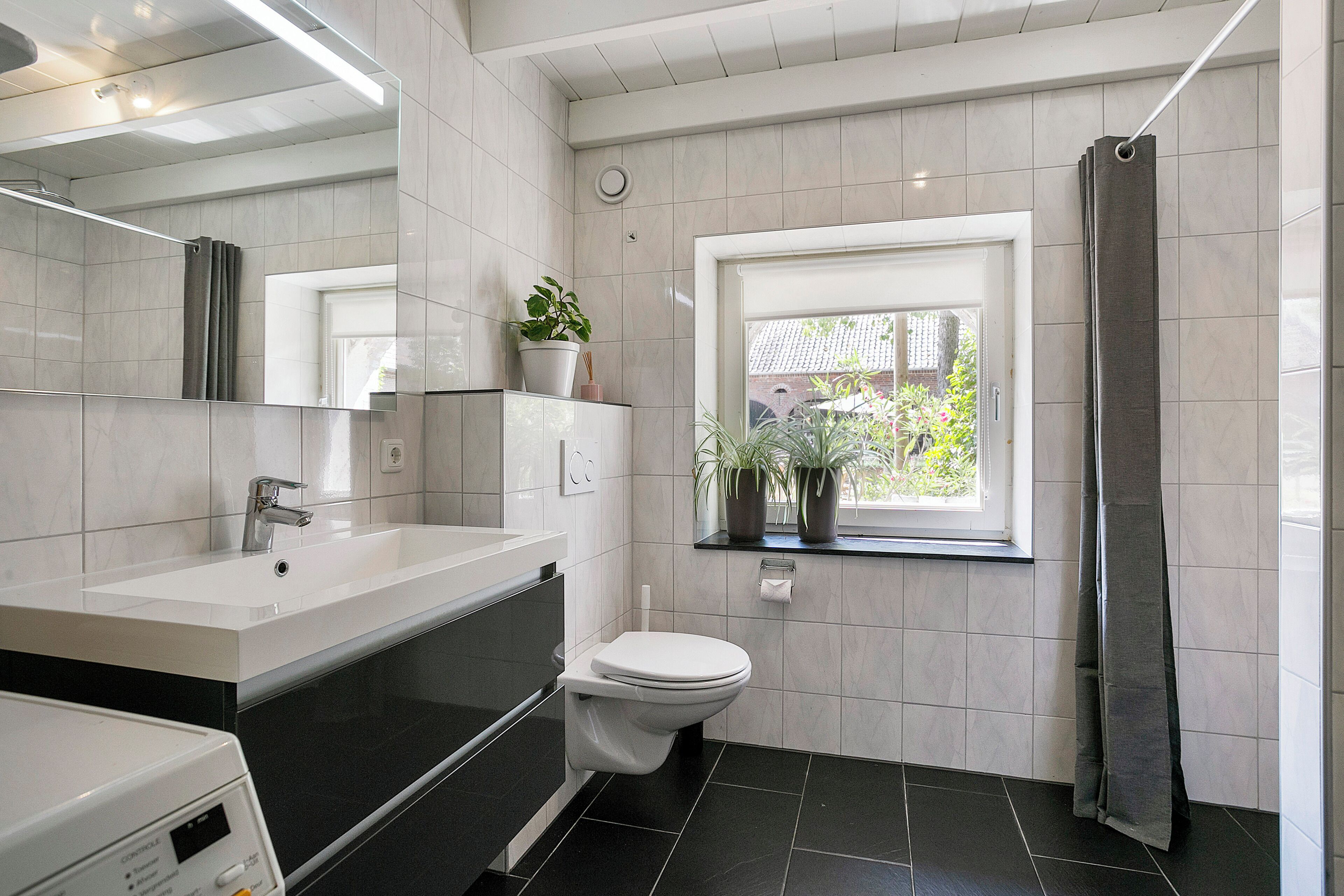 Ein modernes Badezimmer mit Waschbecken in Weiß, schwarzer Arbeitsplatte und Toilette. Ein Fenster bietet Blick auf Grünflächen.