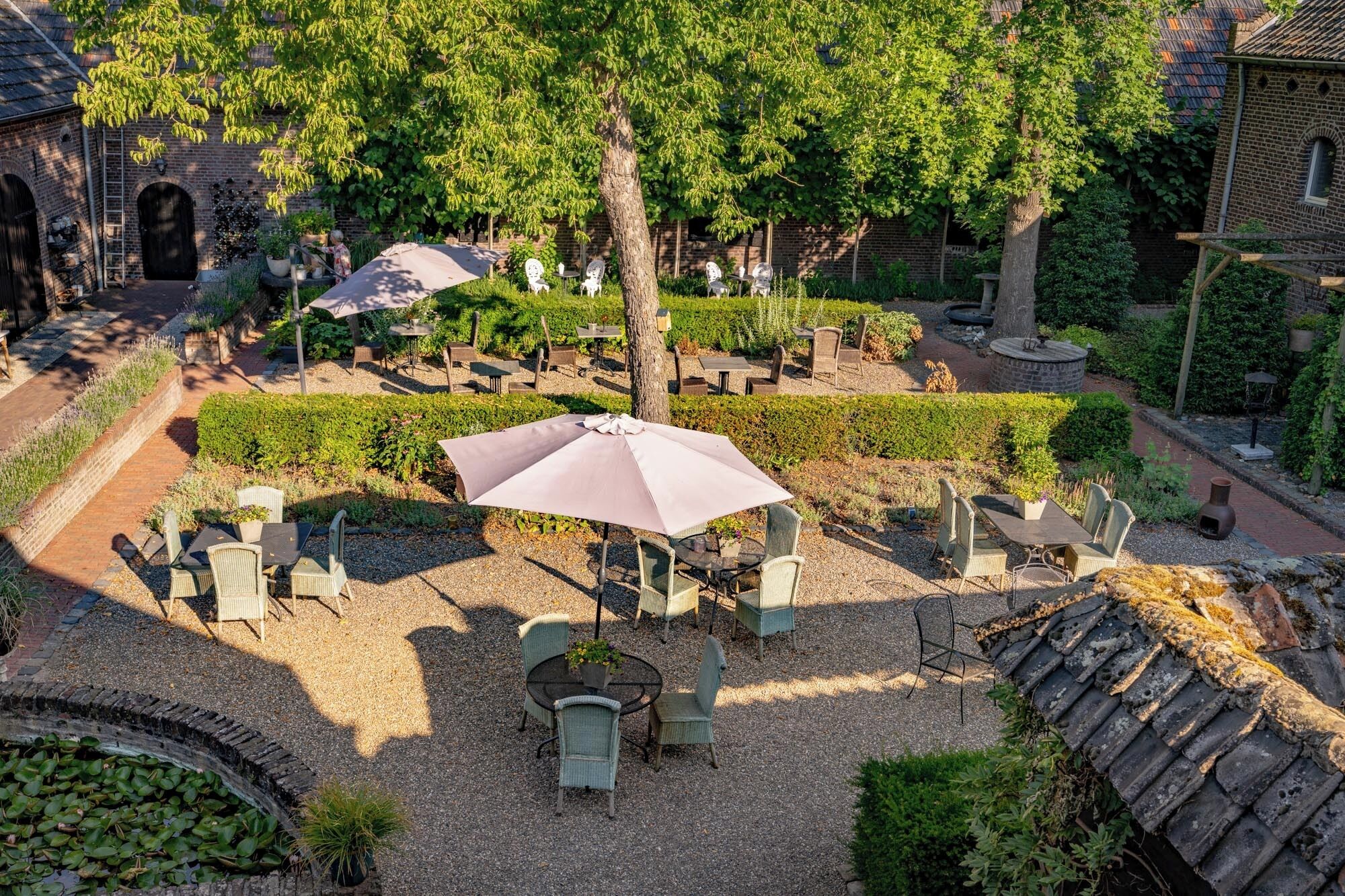 An outdoor patio area with tables and chairs, surrounded by greenery and a brick building.
