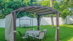 A covered outdoor seating area with a table and chairs, surrounded by a white wall and trees. - mirok gardens kisumu (Kisumu)