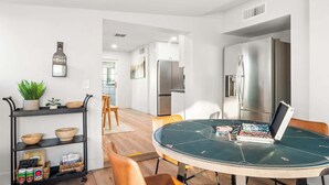 Una cocina moderna con electrodomésticos de acero inoxidable, un comedor con mesa redonda, y una estantería de metal negro con cuencos y una planta.