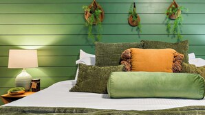 Una cama con ropa de cama verde, varios almohadones, una lámpara de noche y una pequeña planta sobre una mesita de noche de madera.