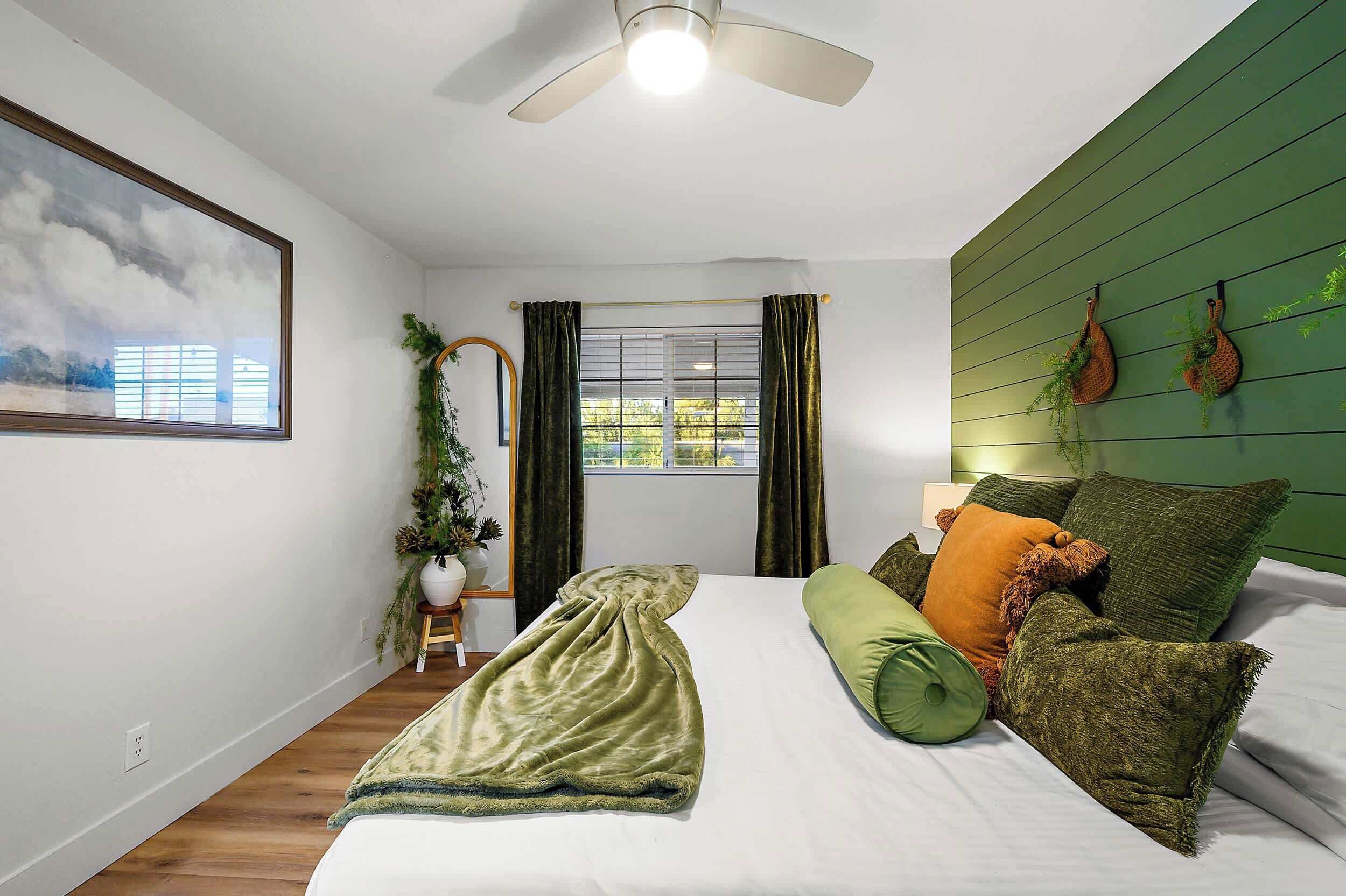 A bedroom with a bed, green walls, a ceiling fan, a window with curtains, and a framed picture on the wall.