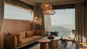 Un salon moderne avec un canapé, deux petites tables rondes en bois, un fauteuil en osier et une vue sur le paysage grâce à de grandes fenêtres.