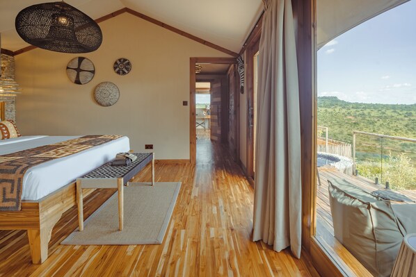 A bedroom with a wooden floor, a bed with a patterned bedspread, a bench, and a view of a balcony with a scenic landscape. - Pearl Mara (Maasai Mara)
