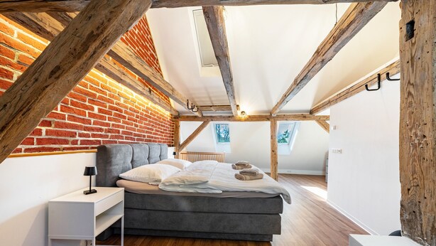 A bedroom with a sloped ceiling, exposed wooden beams, a brick wall, a grey bed, a white bedside table, and a small window.
