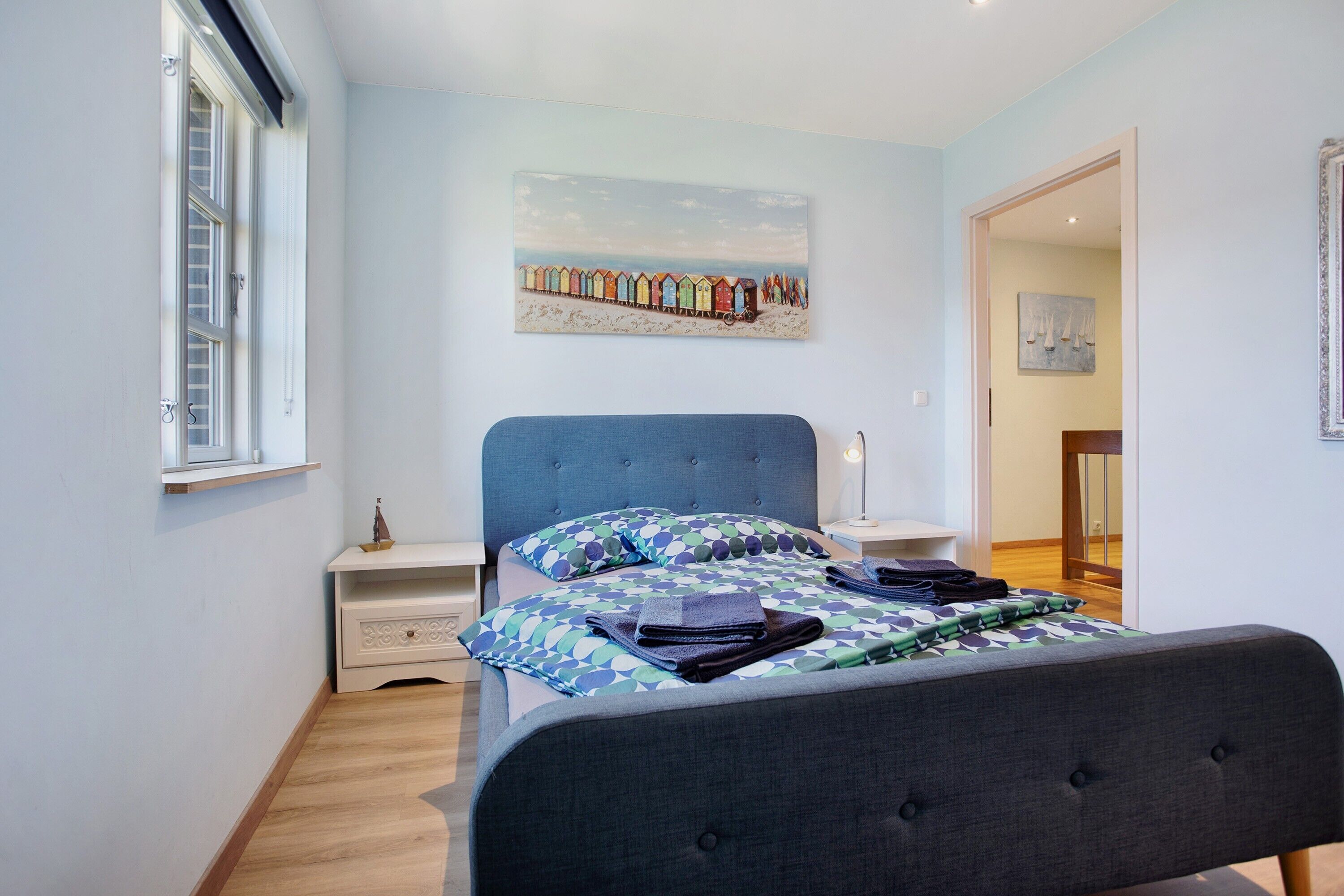 A bedroom with a bed, a nightstand, a window, and a painting on the wall.