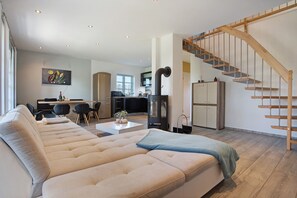 A modern living room with a large beige sectional sofa, a kitchen in the background, and a wooden staircase. - Apartment 'Komfortables Ferienhaus Hafen' with Private Terrace, Private Garden and Wi-Fi (Krummin)