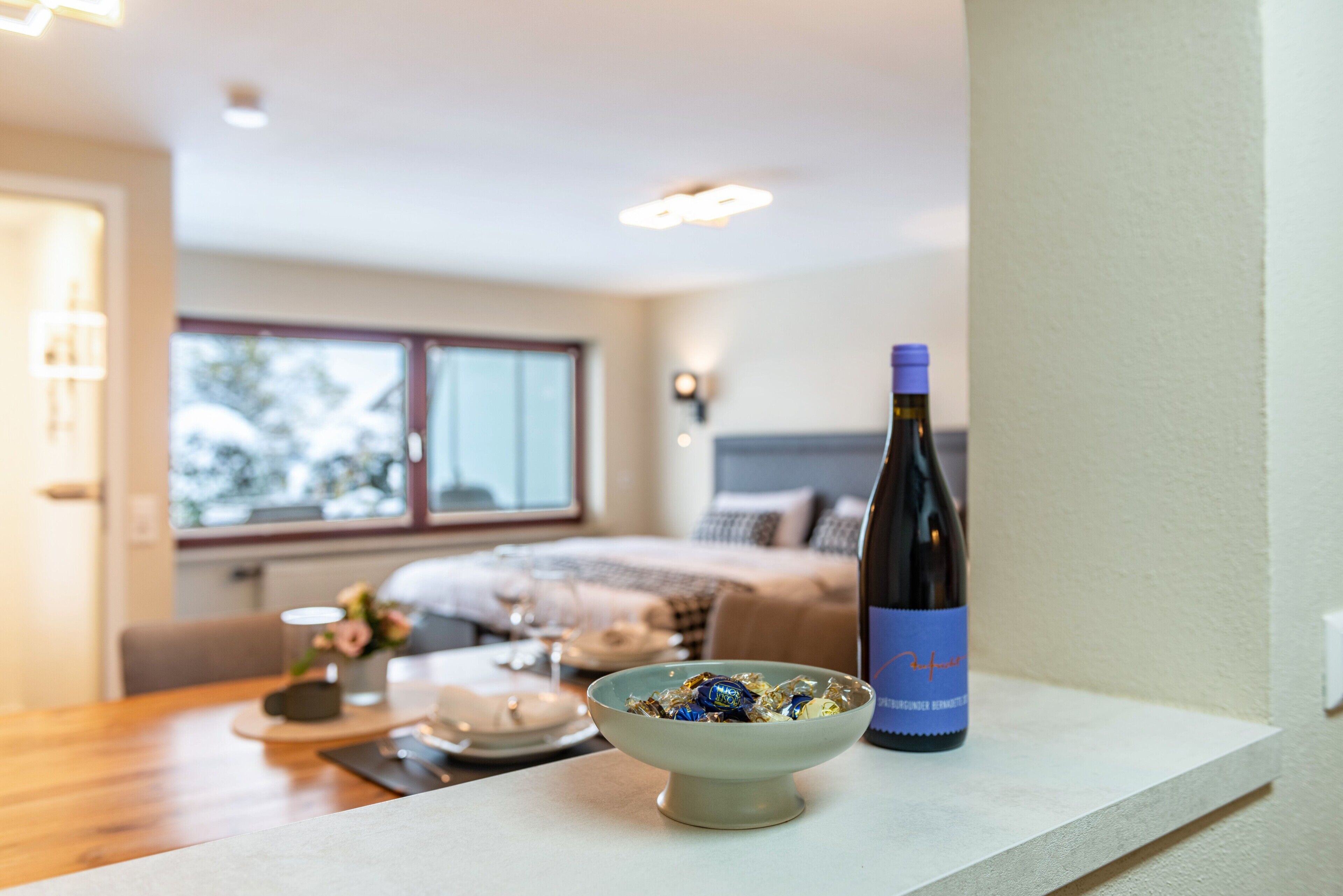 Quarto com cama, mesa de jantar com um recipiente de petiscos, garrafa de vinho e janela com vista.