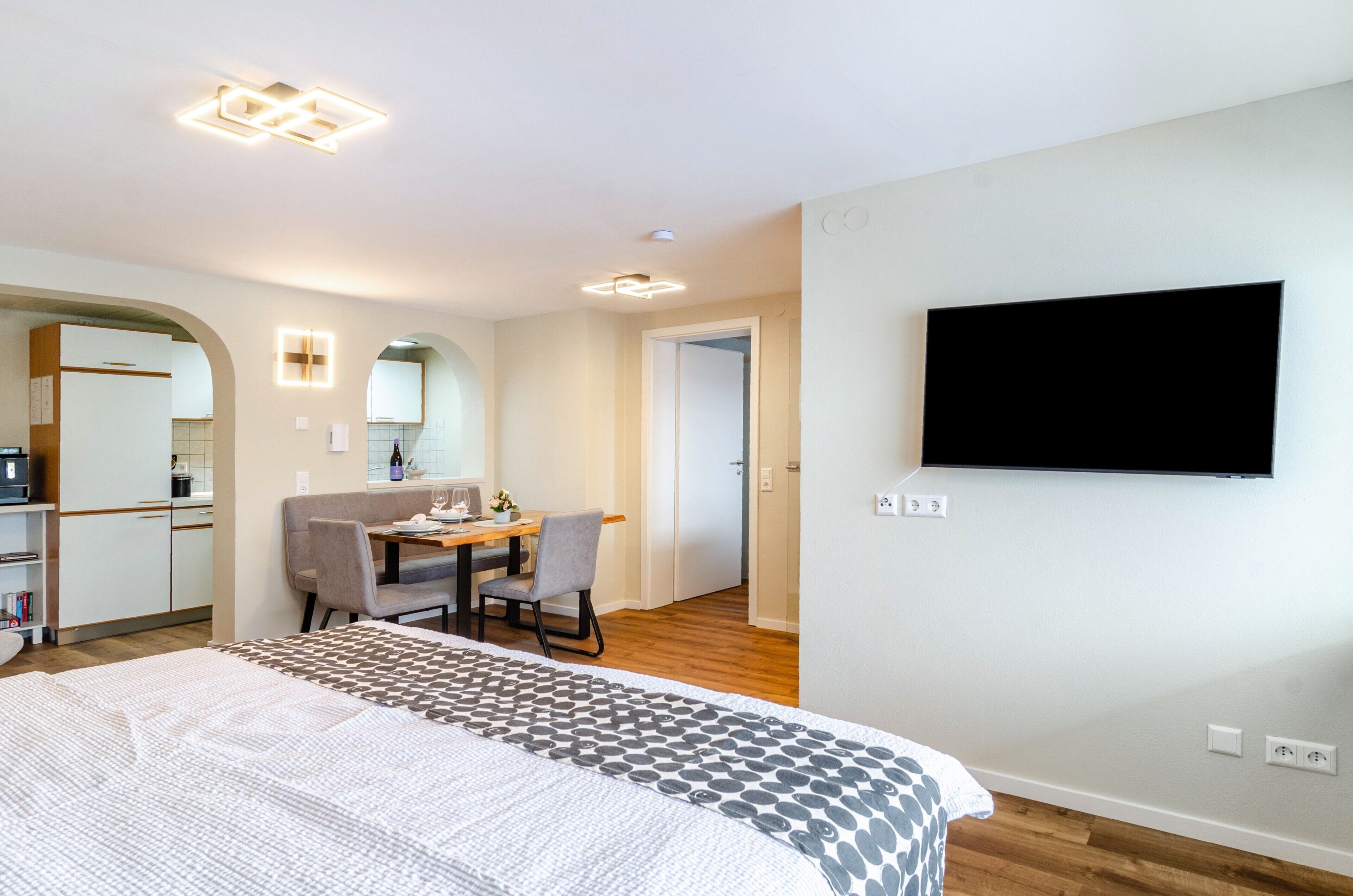 Habitación de hotel con cama, escritorio con silla, televisor montado en la pared y una cocina al fondo.