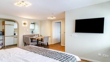Habitación de hotel con cama, escritorio con silla, televisor montado en la pared y una cocina al fondo.