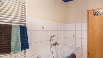 Un bagno con una vasca, un portasciugamani con asciugamani, un soffione e una porta in legno.