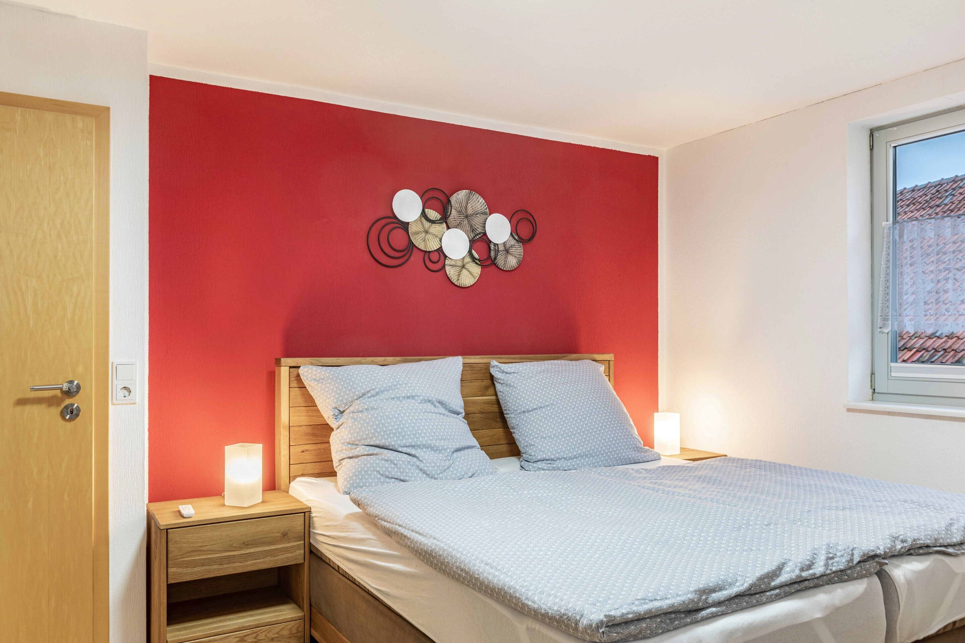 A bedroom with a red wall, a wooden headboard, a bed with white bedding, a bedside table with a lamp, and a window with a view of buildings.