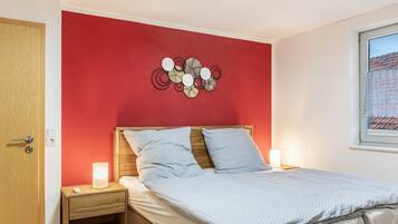 A bedroom with a red wall, a wooden headboard, a bed with white bedding, a bedside table with a lamp, and a window with a view of buildings.