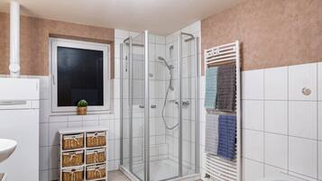 A bathroom with a glass shower enclosure, a white sink, a window with a plant, and a towel rack with multiple towels.