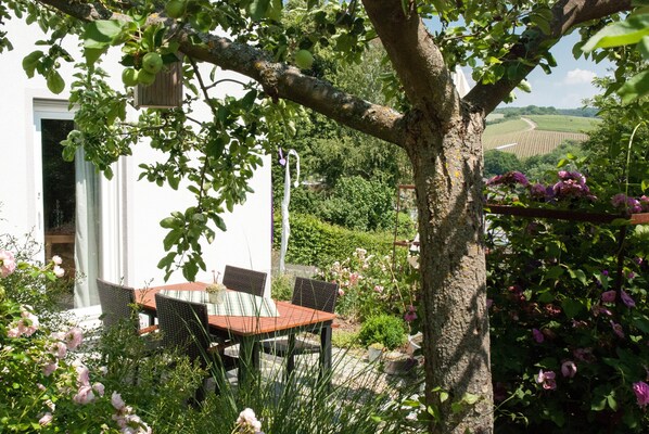 An outdoor dining area with a table and chairs, surrounded by greenery and a tree.