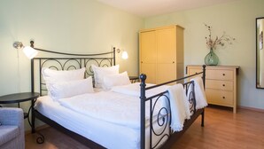 Una cama bien tendida con cabecera, una mesita de noche con lámpara, un aparador y una silla en una habitación con piso de madera.