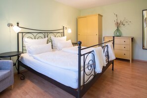 A neatly made bed with a headboard, a bedside table with a lamp, a dresser, and a chair in a room with wooden flooring.