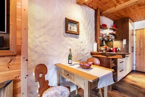 A wooden interior with a dining table, a chair, a bottle of wine, and a bowl of bread.