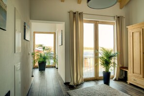 A hallway with a wooden floor, large windows with curtains, and potted plants.
