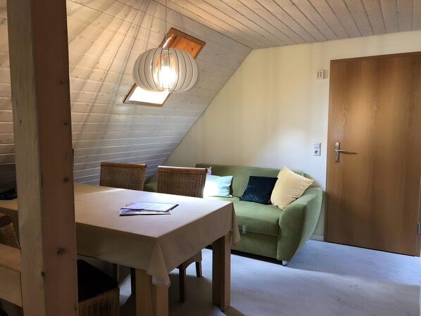 Une pièce comprenant une table à manger, des chaises en osier, un canapé vert, une porte en bois et un plafond en pente avec une lucarne.