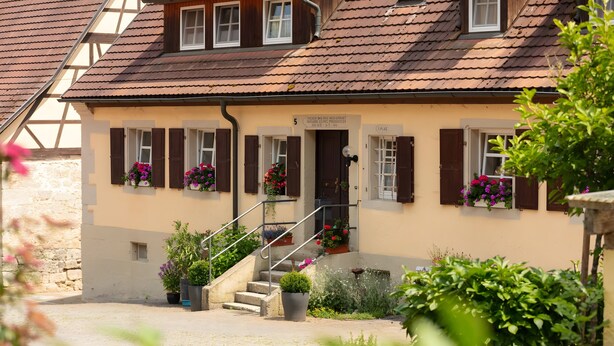 Un bâtiment à deux étages, avec un escalier menant à l’entrée, des plantes en pot et une enseigne au-dessus de la porte.