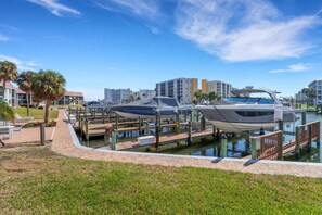 A marina with boats docked, a grassy area with lounge chairs, and buildings in the background. - Beautifully Remodeled Waterfront Condo | Community Pool & Beach Gear | Walk to Santini Plaza & Beach (Fort Myers Beach)