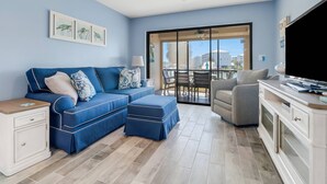 A modern living room with a blue sofa, a grey armchair, a TV, and a balcony with a view of buildings. - Beautifully Remodeled Waterfront Condo | Community Pool & Beach Gear | Walk to Santini Plaza & Beach (Fort Myers Beach)