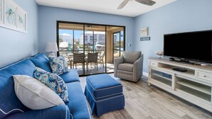 A living room with a blue sofa, a grey armchair, a TV, and a ceiling fan. - Beautifully Remodeled Waterfront Condo | Community Pool & Beach Gear | Walk to Santini Plaza & Beach (Fort Myers Beach)