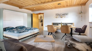 A modern interior with a wooden ceiling, a sofa bed, a dining area with chairs, and a kitchen in the background.
