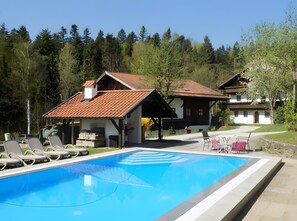 Un espace aménagé au bord de la piscine, avec des chaises longues, une piscine à l’eau d’un bleu limpide et des bâtiments traditionnels en bois surmontés de toits en tuiles rouges.