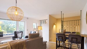 Una sala de estar moderna con sofá, una mesa de comedor con sillas, una zona de cocina y un gran ventanal con vistas a árboles.