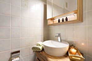 Un baño con un lavabo blanco, un espejo, un estante de madera con artículos de aseo y una toalla.