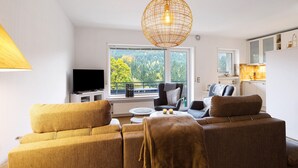 Una sala de estar con sofá, televisión, mesa de comedor y vista a un balcón con árboles.