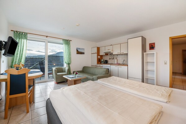 A hotel room with a bed, a desk, a chair, a sofa, a kitchenette, and a view of the mountains.