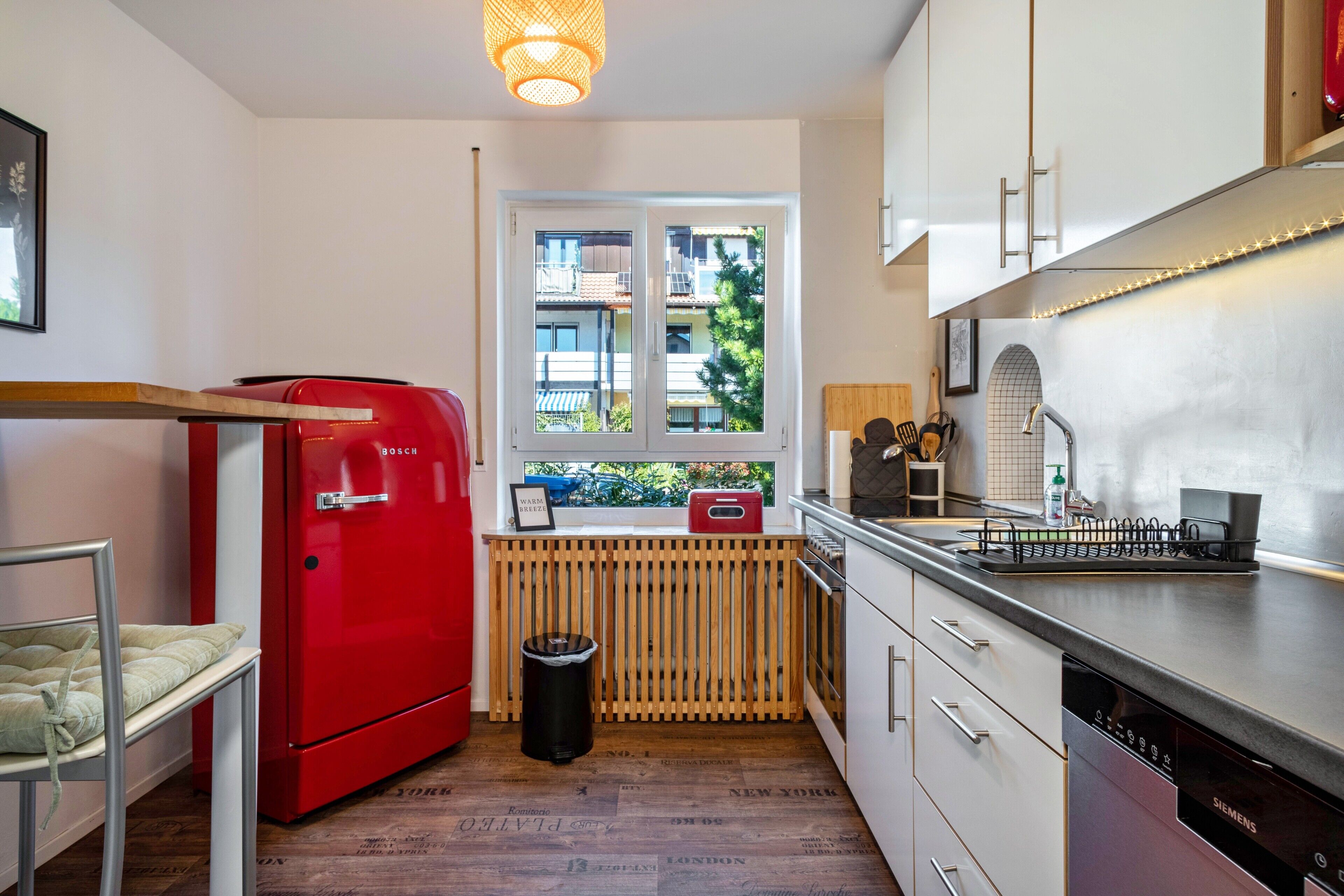 A compact kitchen with a red Bosch refrigerator, a Siemens dishwasher, and a window with a view of buildings.
