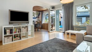 A modern living room with a flat-screen TV, a bookshelf, a sofa, a coffee table, and a large window with curtains. - Casa Noho: Private Terrace, Garden, Wi-Fi – Lake Constance Retreat for Families & Friends (Nonnenhorn)