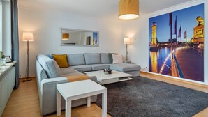 A modern living room with a sectional sofa, a white coffee table, a large painting of a waterfront, and a floor lamp. - Casa Noho: Private Terrace, Garden, Wi-Fi – Lake Constance Retreat for Families & Friends (Nonnenhorn)