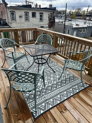 Eine Holzterrasse mit einem Metalltisch und -stühlen, einem gemusterten Teppich und Blick auf Gebäude im Hintergrund.