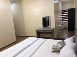 Una habitación de hotel con una cama, una mesita de noche, un espejo y un ventilador de pie.